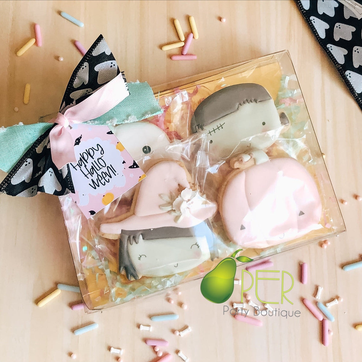 A clear gift box containing four decorated Halloween-themed cookies shaped like ghosts and a skeleton, tied with a pink bow and tag that reads 'Happy Halloween.' The box sits on a light wood surface with scattered pastel sprinkles, and a black-and-white ghost-patterned ribbon is draped at the top right.