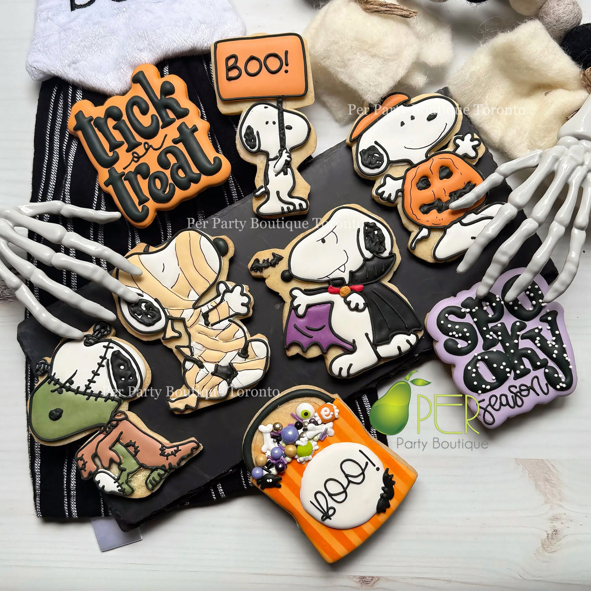 A flat lay of Halloween-themed sugar cookies decorated with Peanuts characters and seasonal motifs, arranged on a black fabric backdrop with skeleton hands and plush props around them. Visible text includes 'Trick or treat', 'Boo!', 'Snoopy', 'Halloween', and 'Spooky Season' on the cookie designs.
