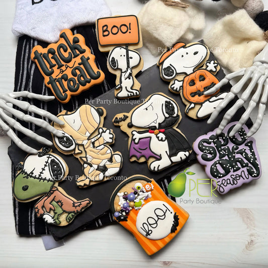 A flat lay of Halloween-themed sugar cookies decorated with Peanuts characters and seasonal motifs, arranged on a black fabric backdrop with skeleton hands and plush props around them. Visible text includes 'Trick or treat', 'Boo!', 'Snoopy', 'Halloween', and 'Spooky Season' on the cookie designs.