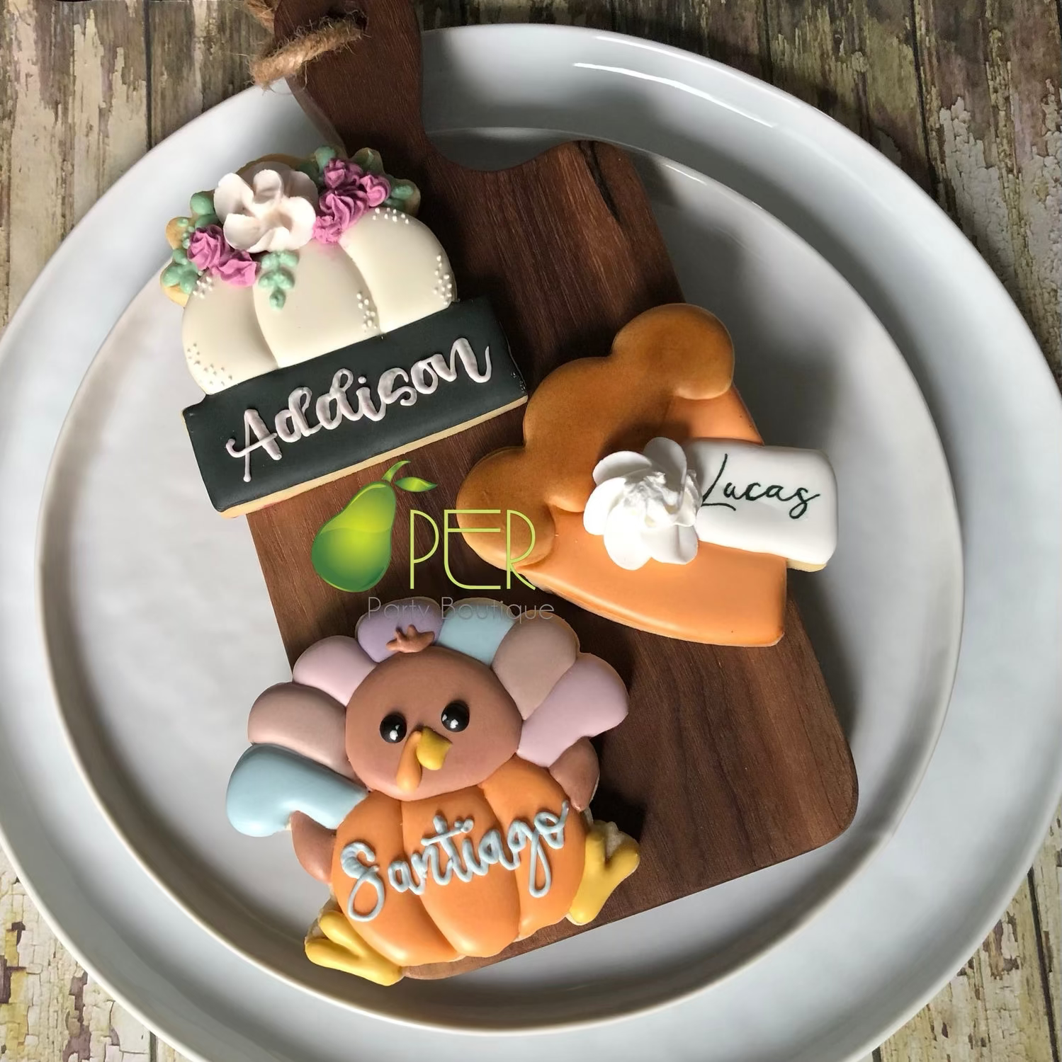 Three personalized sugar cookie cutouts shaped like a pumpkin, a slice of pumpkin pie, and a turkey, arranged on a wooden serving board over a white plate. The names 'Addison', 'Lucas', and 'Santians' are written in icing on the cookies.
