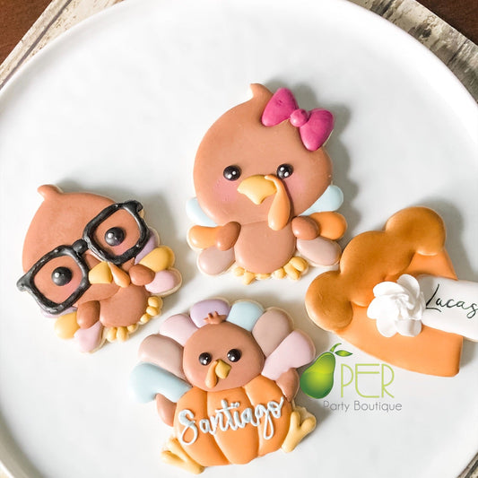 Four decorated sugar cookies shaped like turkeys and a house, arranged on a white plate. The cookies are decorated with pastel orange, peach, pink, yellow, and blue icing, with the names 'Lucas' and 'Santiago' written on them.