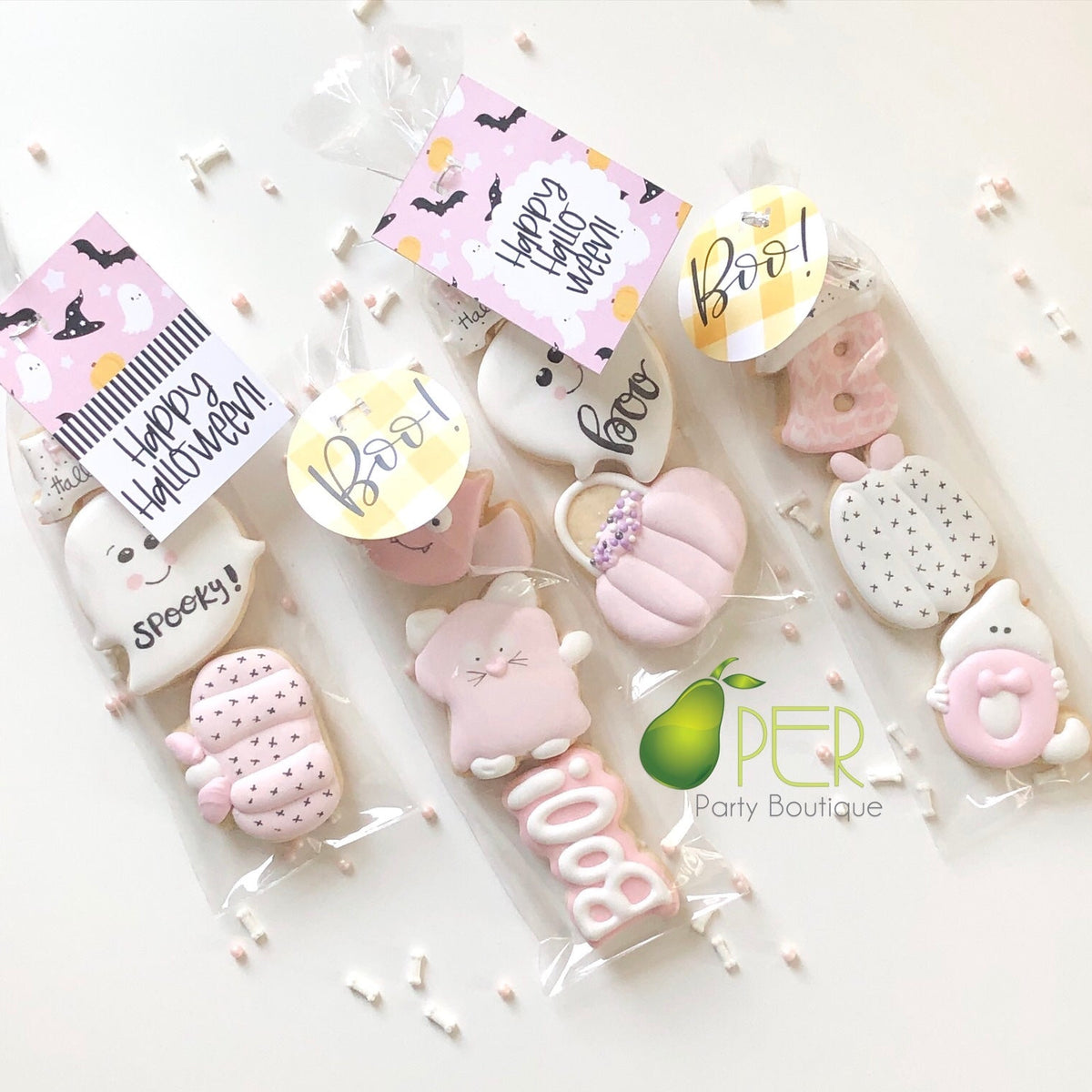 Three Halloween-themed sugar cookies packaged in clear cellophane bags with decorative tags. The cookies are decorated with pink, white, and pastel colors, featuring cute designs like a ghost, cat, pumpkin, spider, and donut.