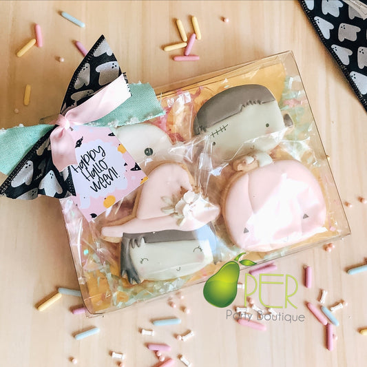 A clear gift box containing four decorated Halloween-themed cookies shaped like ghosts and a skeleton, tied with a pink bow and tag that reads 'Happy Halloween.' The box sits on a light wood surface with scattered pastel sprinkles, and a black-and-white ghost-patterned ribbon is draped at the top right.